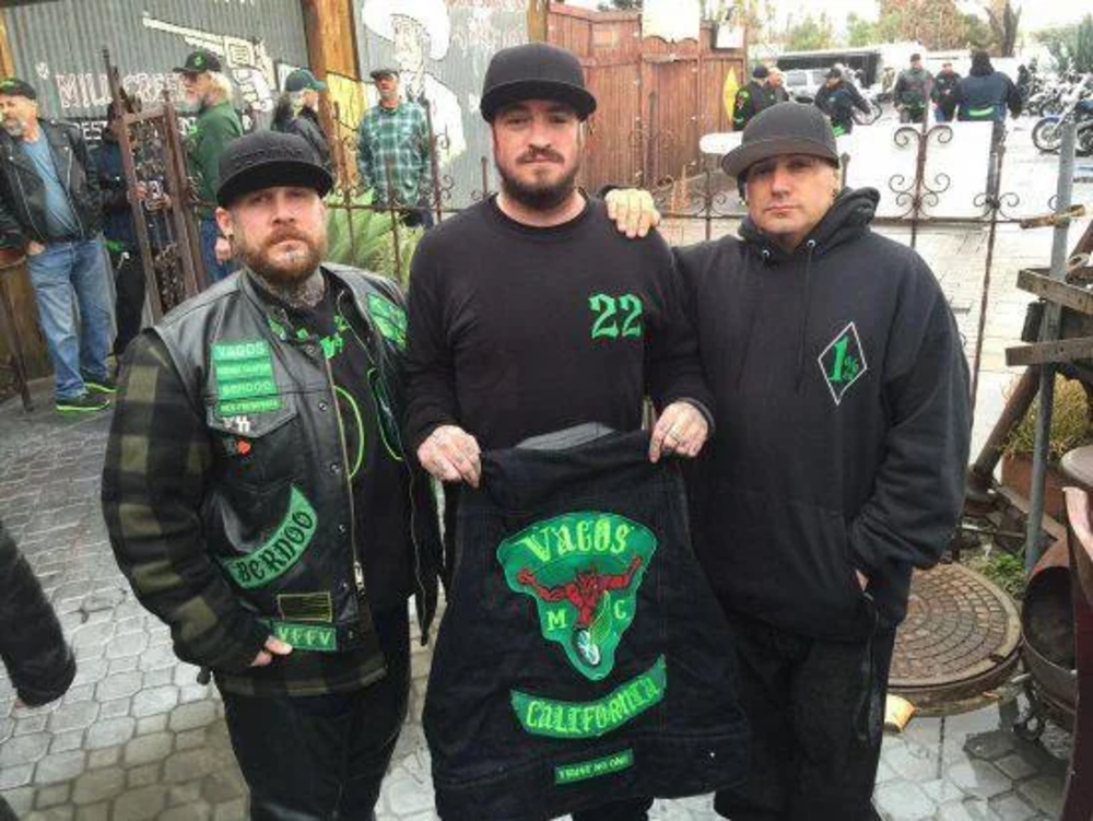 Three men wearing black and green clothing stand outdoors. The man in the center holds a jacket with "Vagos MC" and a design featuring a red bull's skull with green horns. Other people and motorcycles are visible in the background.