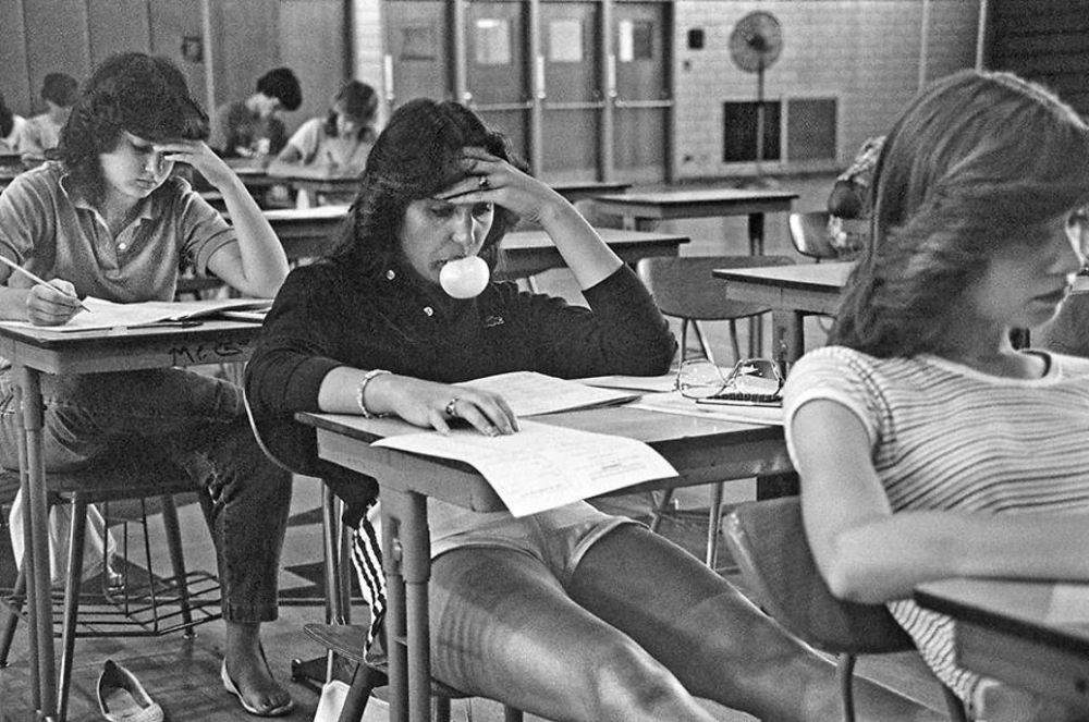 Black and white photo of students sitting at desks in a classroom, taking a test. A girl in the center is blowing a bubblegum bubble while reading her paper. Other students are focused on their work, with some writing and one resting on the desk.