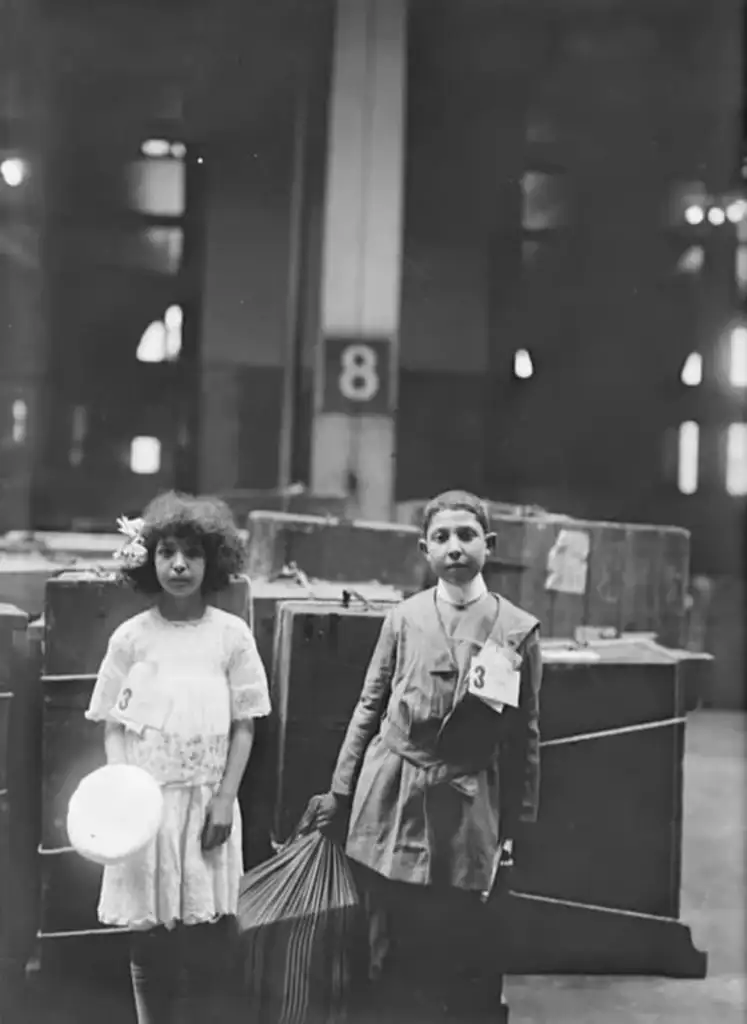 Two children stand in a large immigration processing area. The girl on the left wears a white dress, and the boy on the right wears a dark suit. Both have identification tags. Large crates and a pillar marked with the number 8 are in the background.