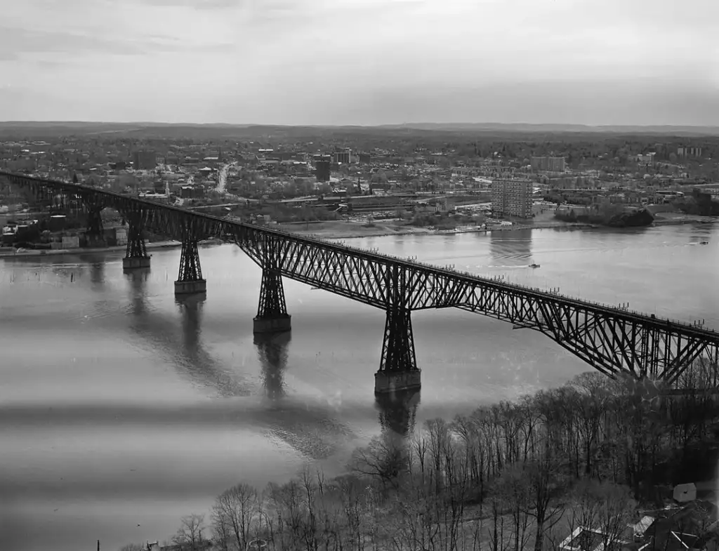 A black and white aerial view of a large iron bridge spanning a wide river. The bridge has a lattice structure with several support piers in the water. In the background, a cityscape is visible under a cloudy sky. Trees line the riverbank in the foreground.