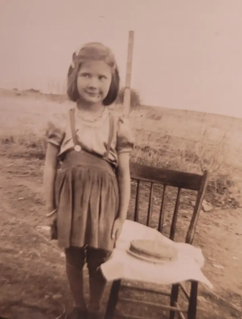 A vintage photo of a young girl with short hair, wearing a dress and standing in front of a wooden chair outdoors. The chair has a cloth with a round loaf of bread on it. The background is an open field with a post.