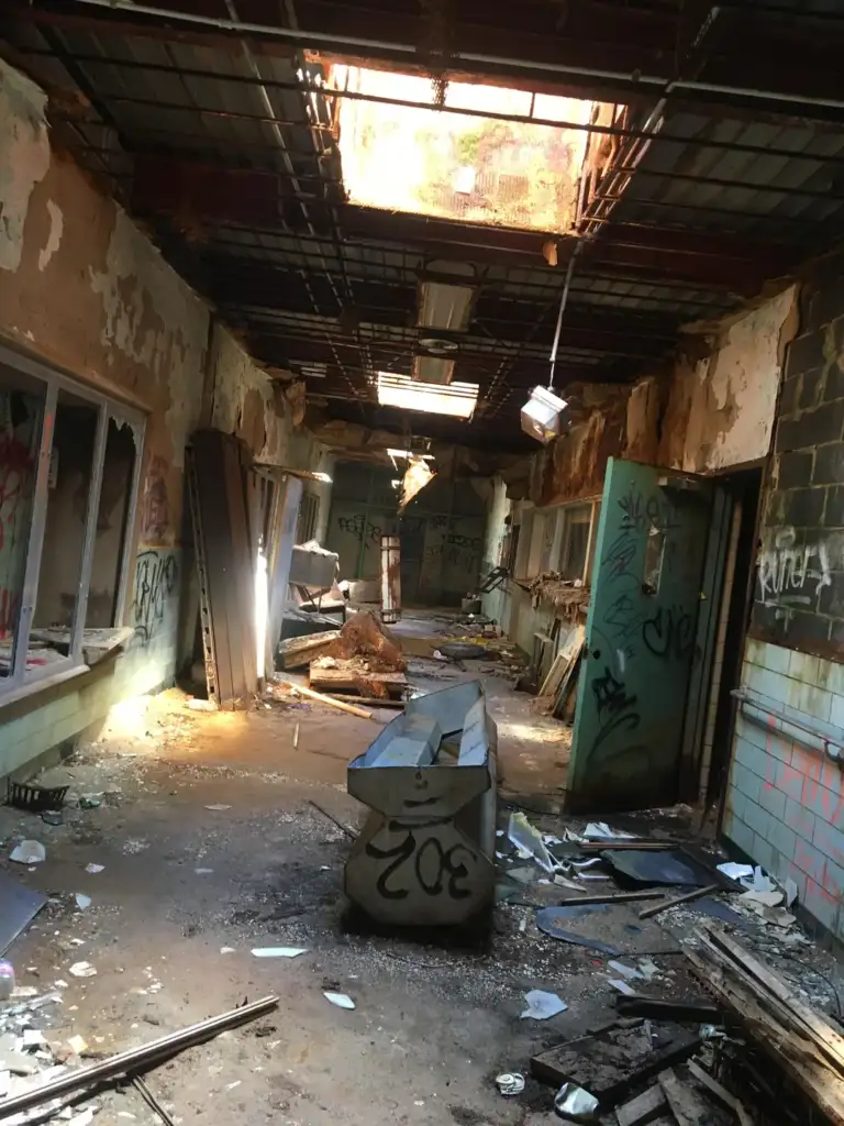 Abandoned narrow hallway with a partially collapsed ceiling, exposing sunlight. Walls are covered in graffiti, debris scattered across the floor, broken windows, and an overturned metal object in the center. Peeling paint and rubble add to the decay.