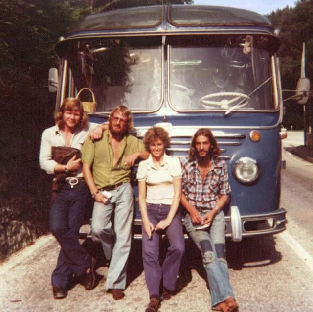 Four people are posing in front of a vintage blue bus on a road. One person is leaning against the bus, while two others are sitting on its bumper. The fourth stands slightly apart. Trees line the background.