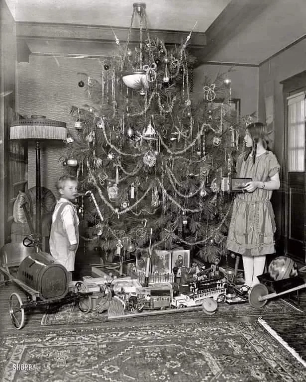 A vintage black and white photo of two children by a decorated Christmas tree. The boy stands next to a toy car, and the girl holds a box. Various toys, including a train set, are placed near the tree.