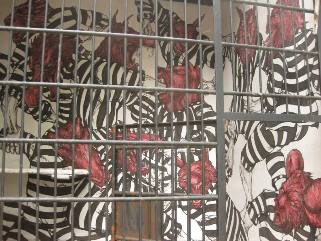 A mural depicting chaotic figures with red heads and striped clothing behind bars. The artwork is complex and detailed, conveying a sense of confinement and movement within a confined space.