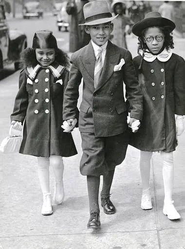 A young boy and two girls, all dressed in formal 1940s attire, walk hand in hand on a sidewalk. The boy wears a suit and hat, while the girls wear coats with matching hats. A woman and a car are visible in the background.