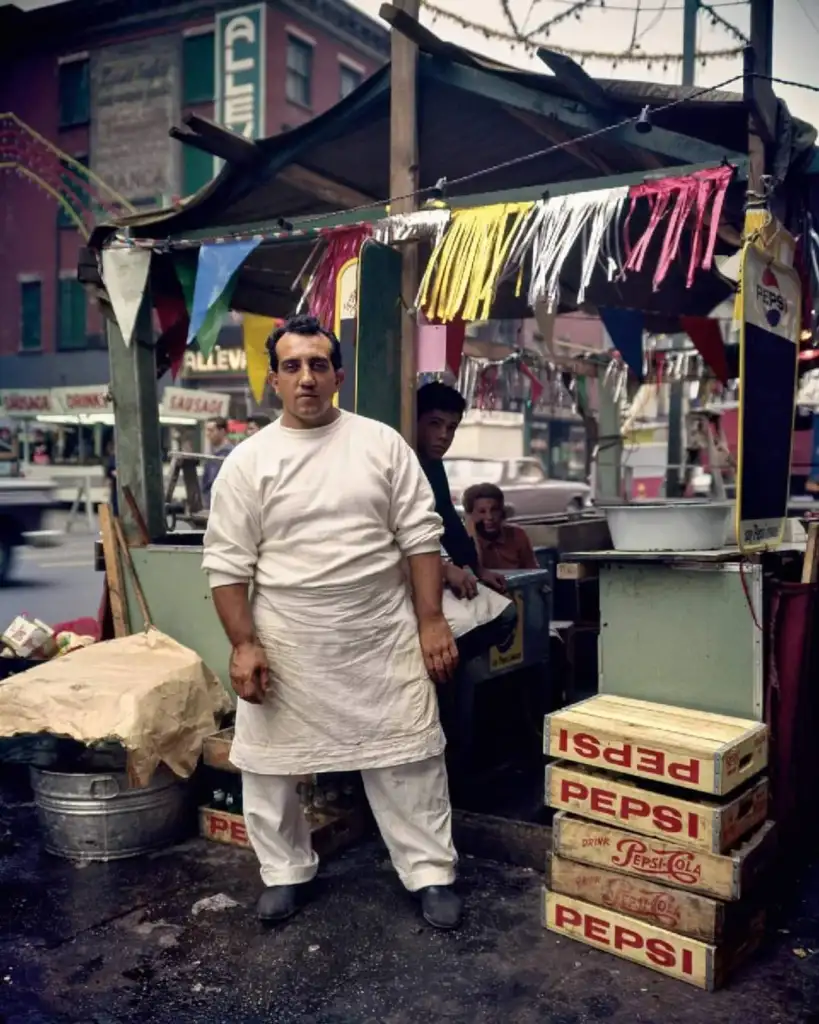 A man in a white chef's outfit stands outside a small food stand decorated with colorful pennants. The stand has Pepsi signage and is set on a street. Two other people are seated inside. Buildings and traffic are visible in the background.