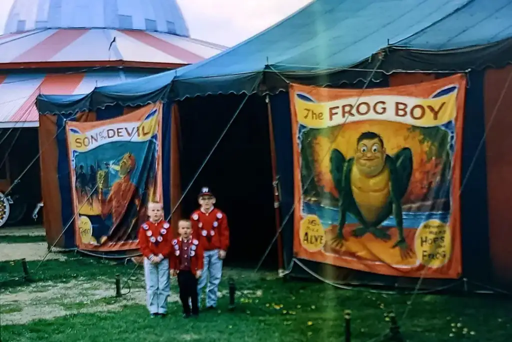 Three children in red jackets stand outside a circus tent with vintage posters. One poster features "Son of Devil" and the other "The Frog Boy." A red and white striped tent is partially visible in the background.