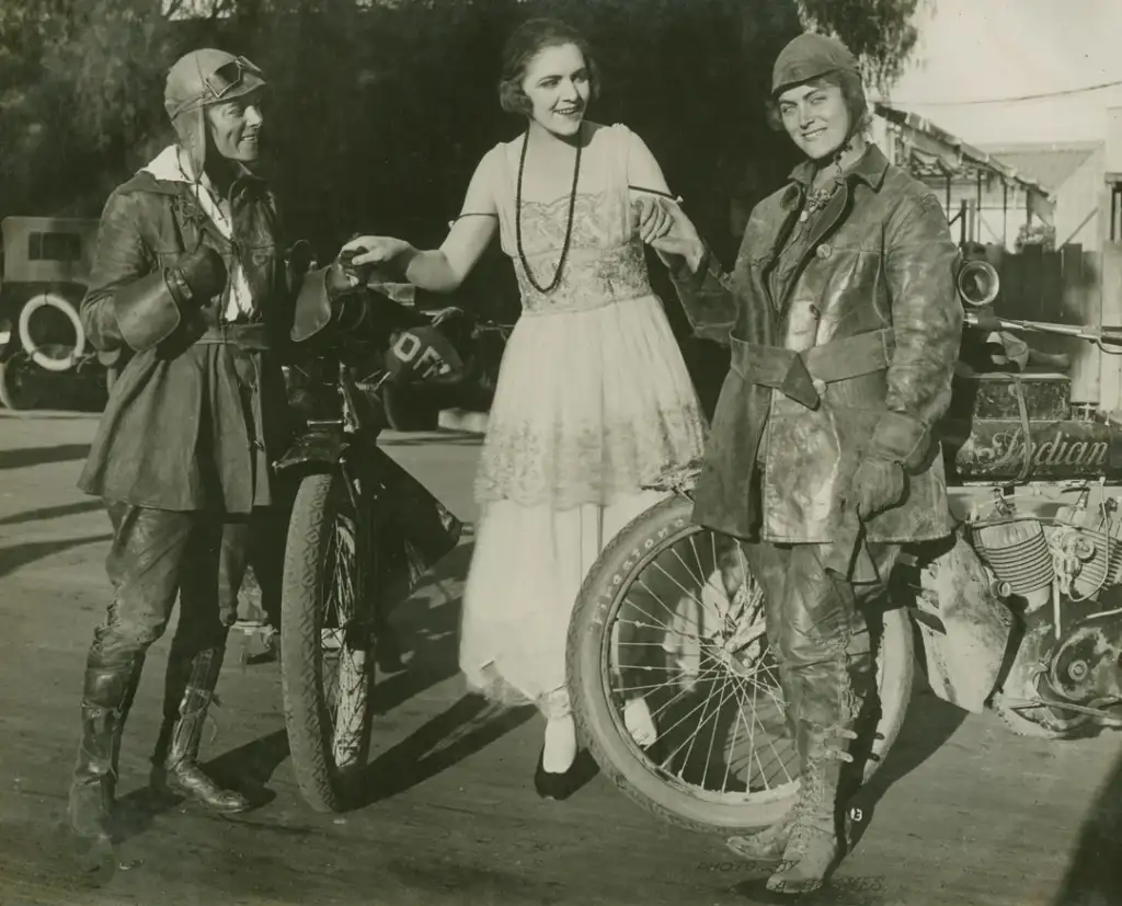 Three women pose with vintage motorcycles. Two wear leather riding gear and boots, while the third stands between them in a lace dress and pearls. The image has an old-fashioned, sepia tone, with motorcycles marked "Indian" in the background.