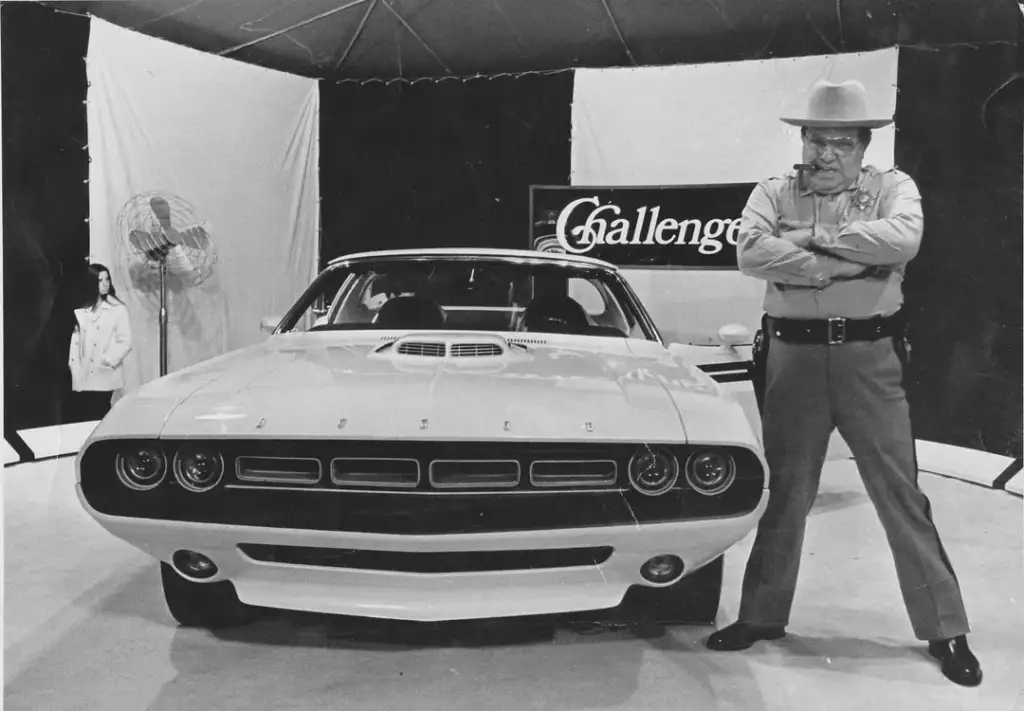 A vintage black and white photo shows a man in a hat and uniform standing proudly beside a classic white muscle car. The car has a prominent grille and "Challenger" sign in the background. A second person and a fan are visible to the left.