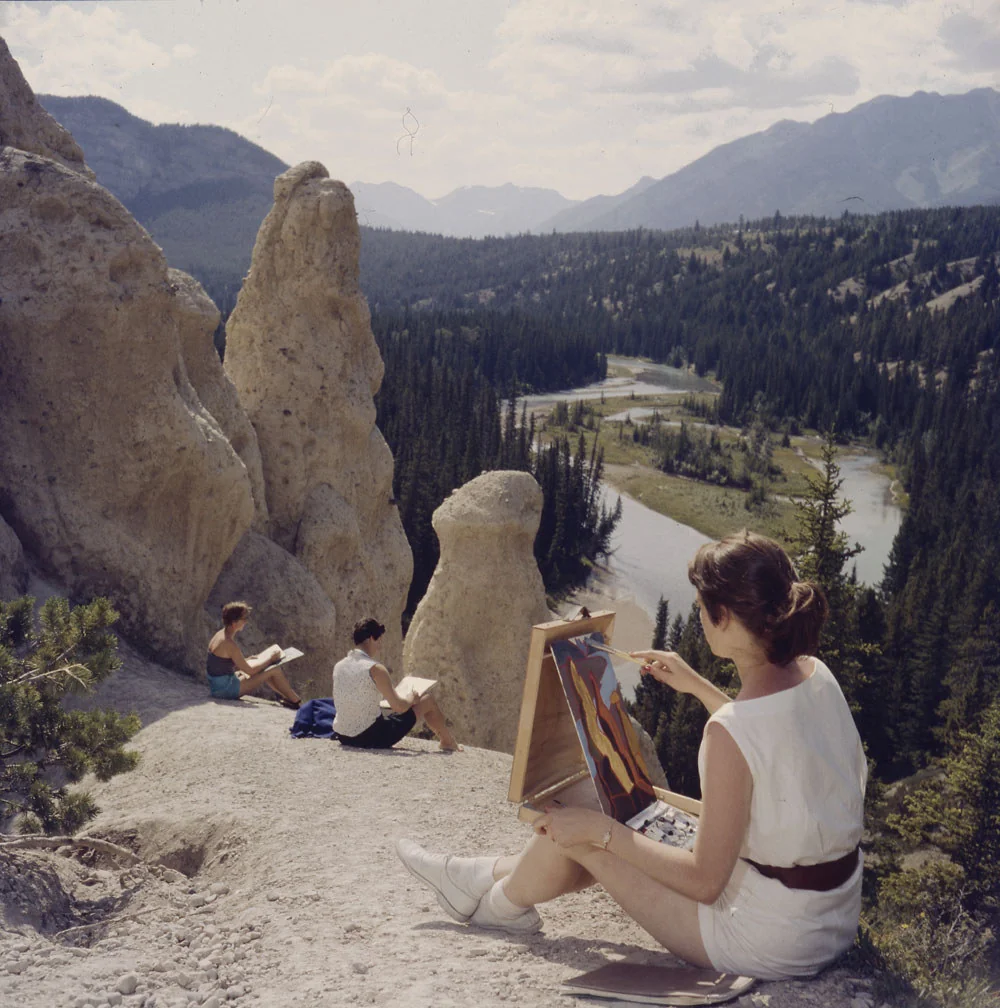 Three people sit on a rocky ledge overlooking a river and forested mountains. One person is painting on an easel, capturing the natural scenery. The sky is partly cloudy, and the view is expansive and serene.