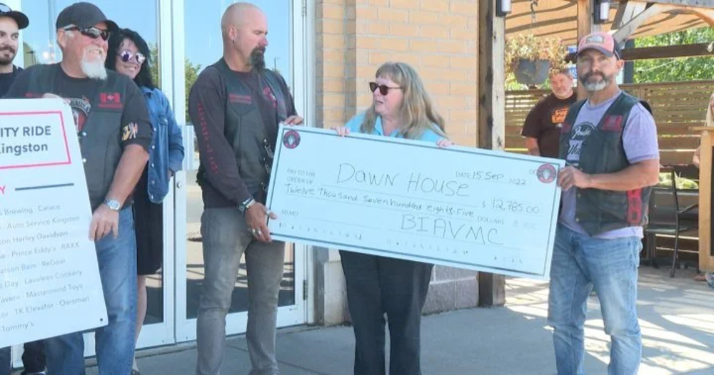 A group of people stand outside holding a large check made out to "Dawn House" for $12,785. One man wears a hat and sunglasses, another has his arm around a woman in sunglasses. They are all smiling.