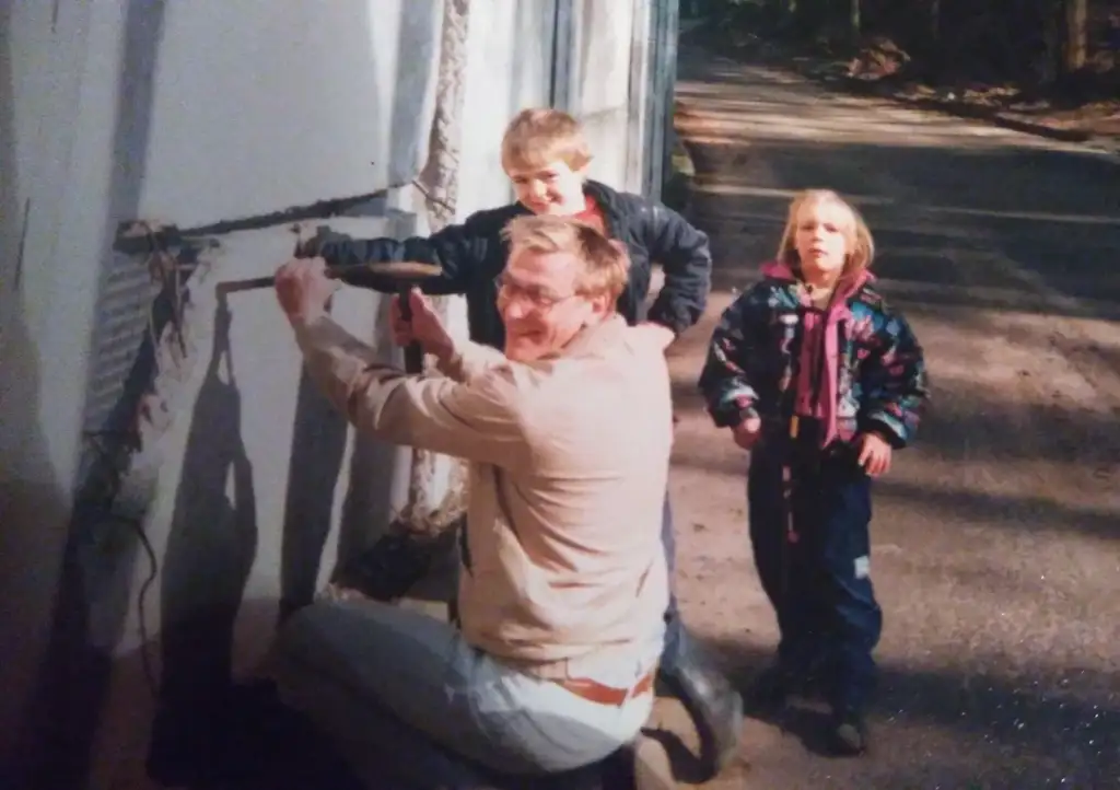 A man kneels while using a hammer on a wall outside. Two children stand nearby on a paved path, watching him. The boy wears a black jacket, and the girl is in a colorful coat. They appear to be in a wooded area.