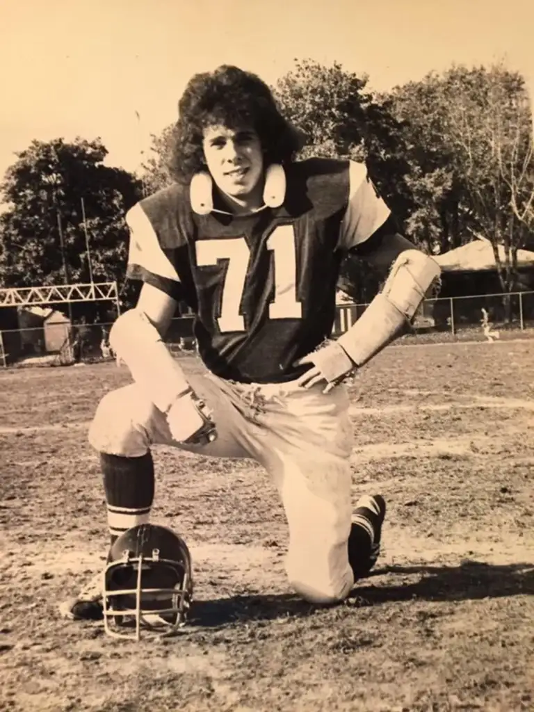 A vintage black and white photo of a football player wearing a jersey with the number 71. He's kneeling on one knee on a football field, surrounded by trees and structures, with a helmet placed on the ground in front of him.