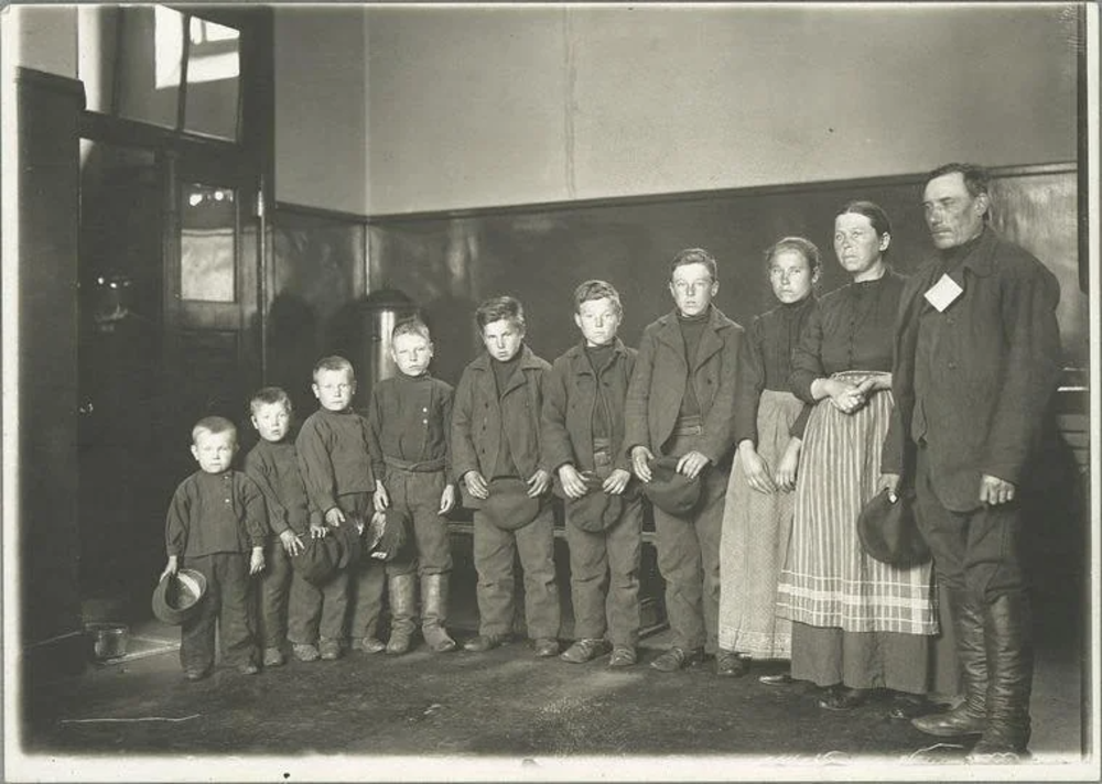 A black and white vintage photo shows a family of ten standing in a line. The group includes eight children holding hats, and two adults, a woman in a plaid skirt and a man wearing a hat. They are in an interior space with plain walls and a door.