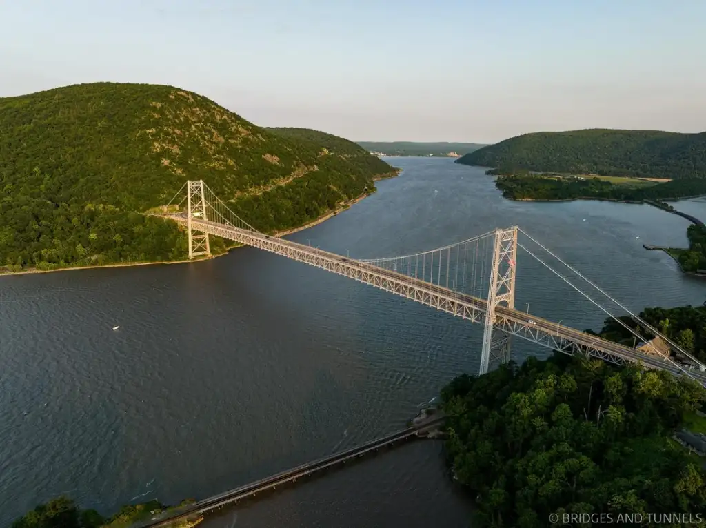 Aerial view of a suspension bridge spanning a wide river, surrounded by lush green hills. The water reflects the clear sky, and a small boat is visible near the left bank. The landscape is serene, with a few roads cutting through the greenery.