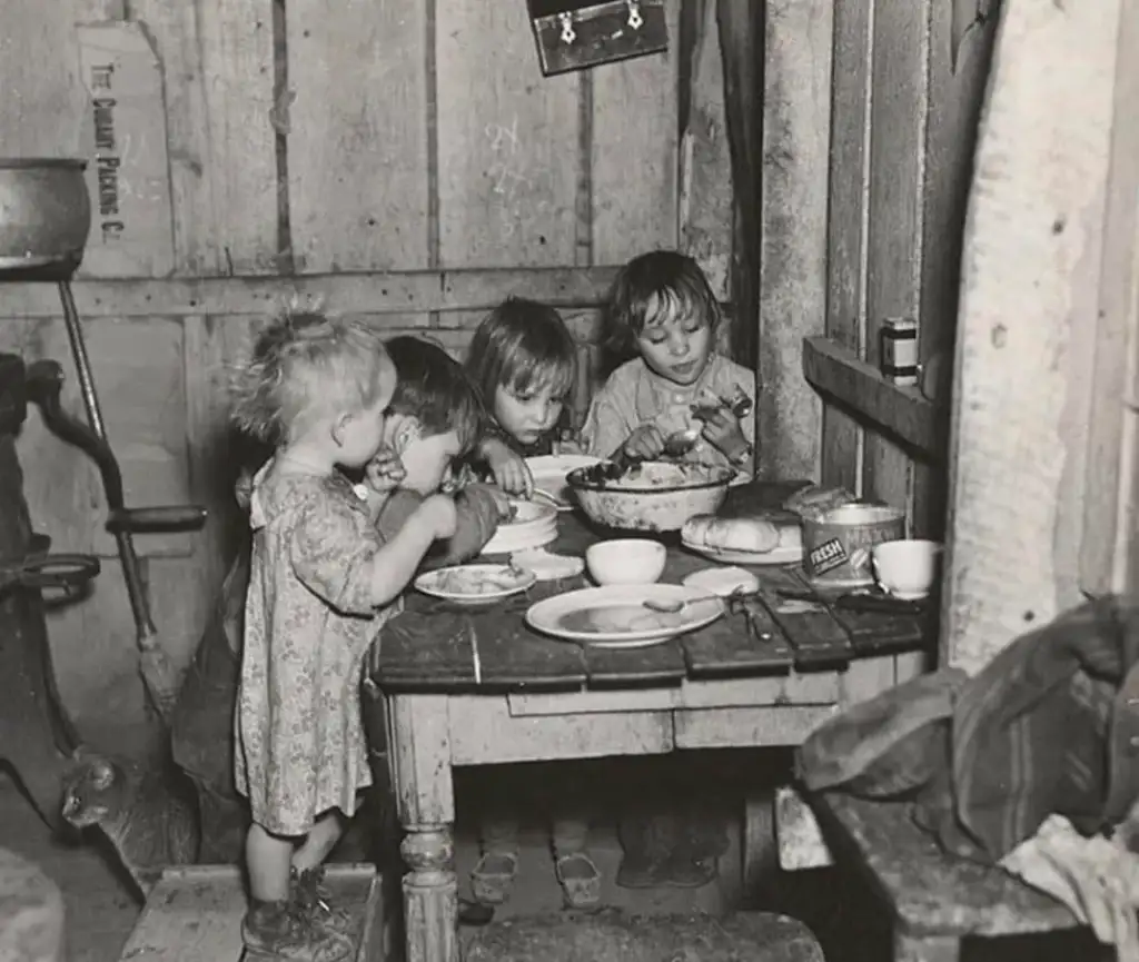 Four young children sit around a rustic wooden table in a dimly lit room, eating from a shared bowl. The table holds simple dishware and a loaf of bread. The walls are wooden and bare, with a grinder visible in the background.