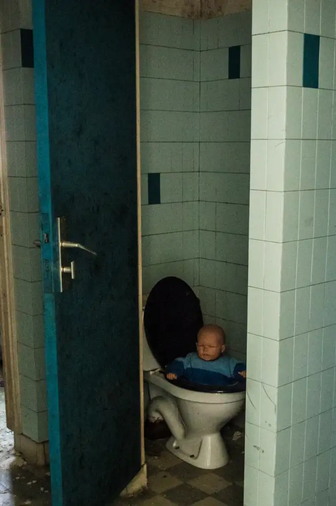 A baby doll is placed inside a toilet bowl with its arms resting on the seat in a small, dimly lit bathroom. The room has light blue tiled walls and a partially open door. The floor is checkered with black and white tiles.