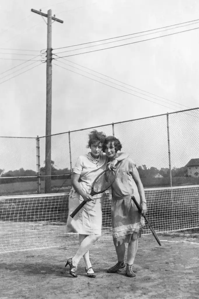 Two women in vintage dresses stand on a tennis court, each holding a racket. They are smiling and leaning toward each other. A chain-link fence and utility pole are visible in the background.