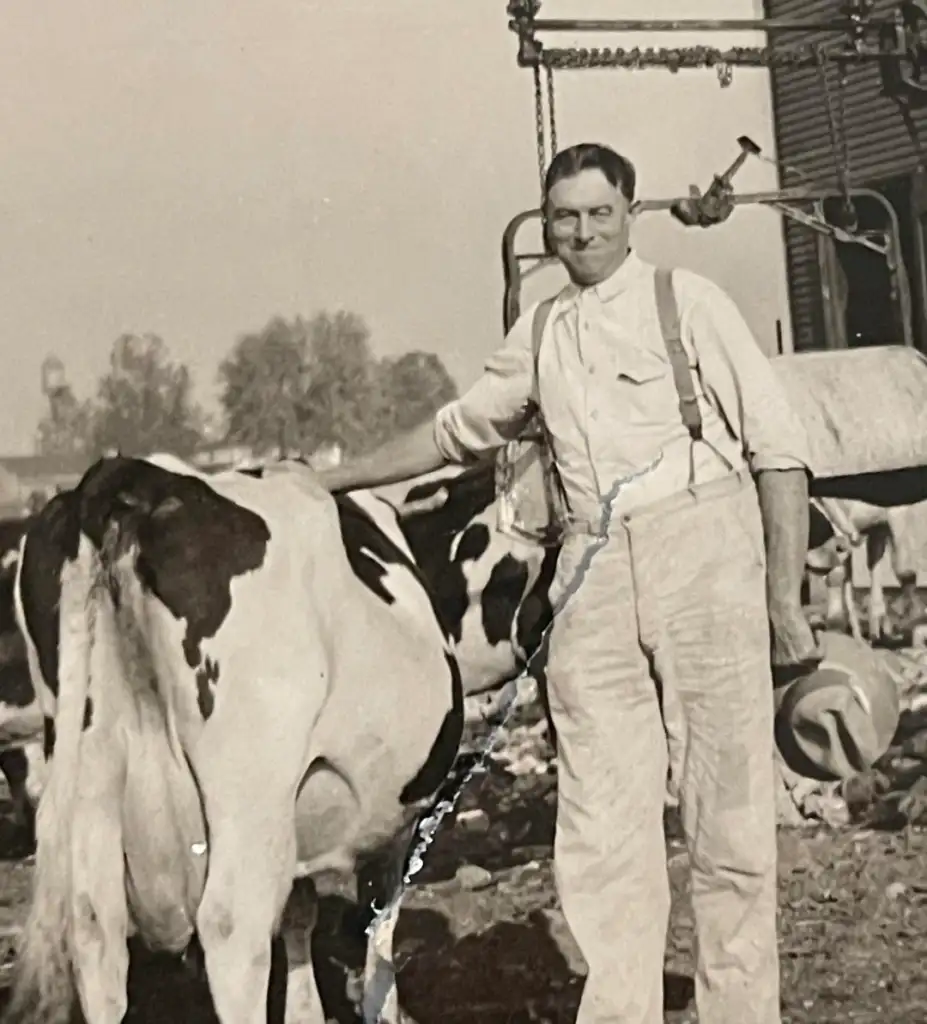 A vintage black-and-white photo of a person in white overalls standing beside a dairy cow, smiling. They hold a hat in one hand. In the background, there is farm equipment and blurred trees.