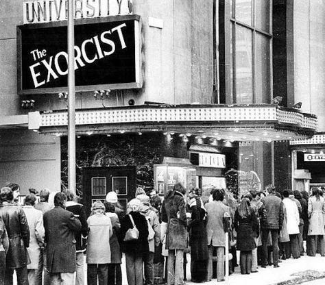 Black and white photo of a long line of people waiting outside a theater. The marquee shows the movie title "The Exorcist." The crowd appears bundled up, suggesting cold weather. The theater displays vintage architecture and signage.