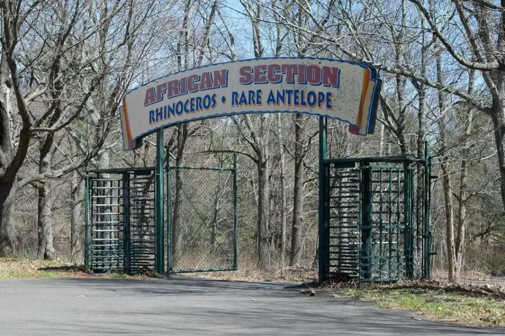 Entrance gate to the "African Section" of a wildlife park, featuring signs for rhinoceros and rare antelope. The area is surrounded by leafless trees, and the path is paved.