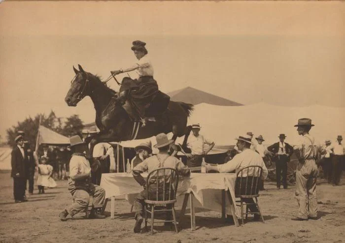 A horse and rider elegantly jump over a dining table surrounded by seated men in a sepia-toned outdoor setting. Tents and onlookers are visible in the background, capturing a historical scene of equestrian skill.