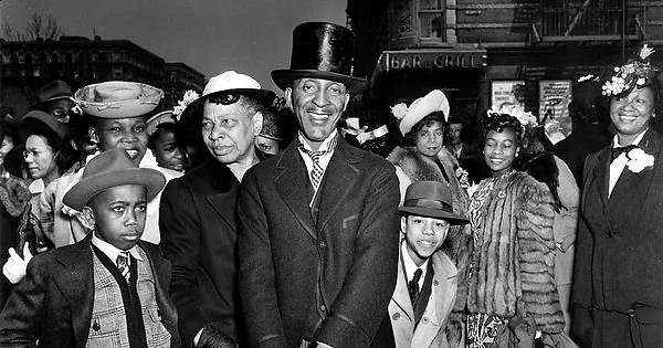 A historic black-and-white photo shows a group of people dressed in elegant attire, including hats and coats, gathered on a street. An older gentleman in a top hat stands in the center, surrounded by smiling adults and children.