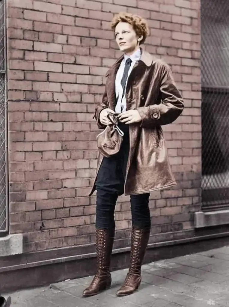 A person stands in front of a brick wall, wearing a long leather coat, white shirt, and tie, holding a small bag in both hands. They are wearing high lace-up boots and have short, curly hair.