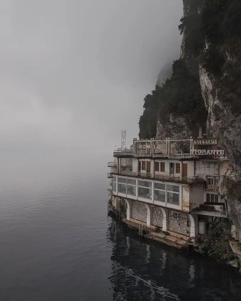 A misty scene shows an abandoned, multi-level building built into a cliffside, overlooking a calm body of water. The structure has a weathered appearance with rust and chipped paint. Dense fog obscures the background, creating a mysterious atmosphere.