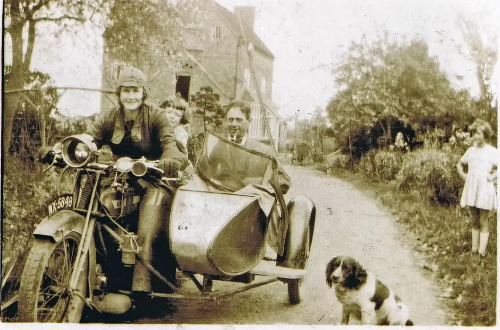 A vintage photograph shows a smiling woman sitting on a motorcycle with a sidecar, holding a child. A man is seated in the sidecar. A dog sits nearby on the dirt road, and a girl stands in the background near a house surrounded by trees.