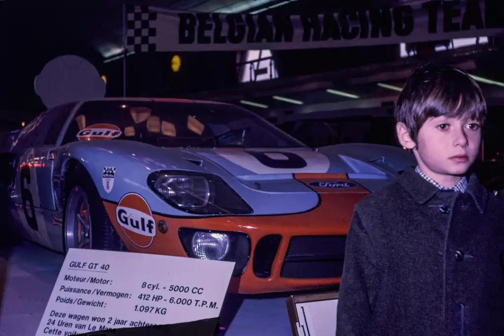 A young boy stands in front of a Ford GT40 sports car with a Gulf livery. The backdrop displays a sign reading "BELGIAN RACING TEAM." Information about the car is visible on a board beside it.