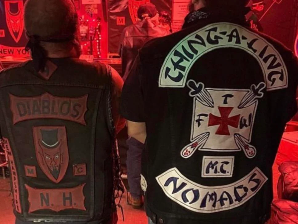 Two people wearing black vests with large patches stand with their backs to the camera. One vest reads "DIABLOS N.H." with a red devil design, and the other reads "THING-A-LING NOMADS" with additional smaller patches.