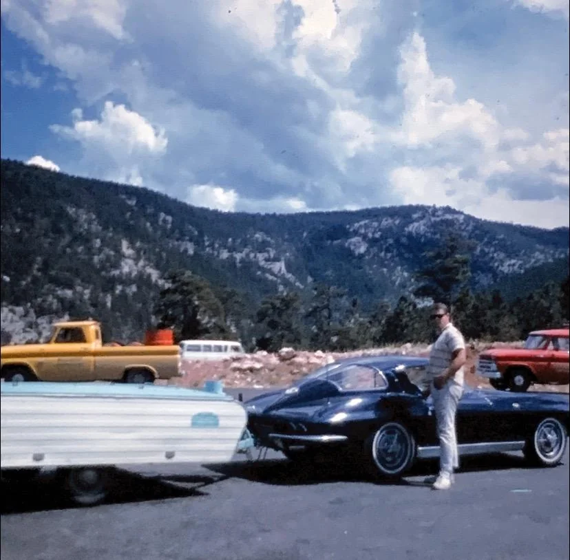 A person stands beside a dark convertible sports car, which is parked on a scenic mountain road. The car is hitched to a small camper trailer. In the background, pine-covered hills and a cloudy sky are visible. Other vehicles are parked nearby.