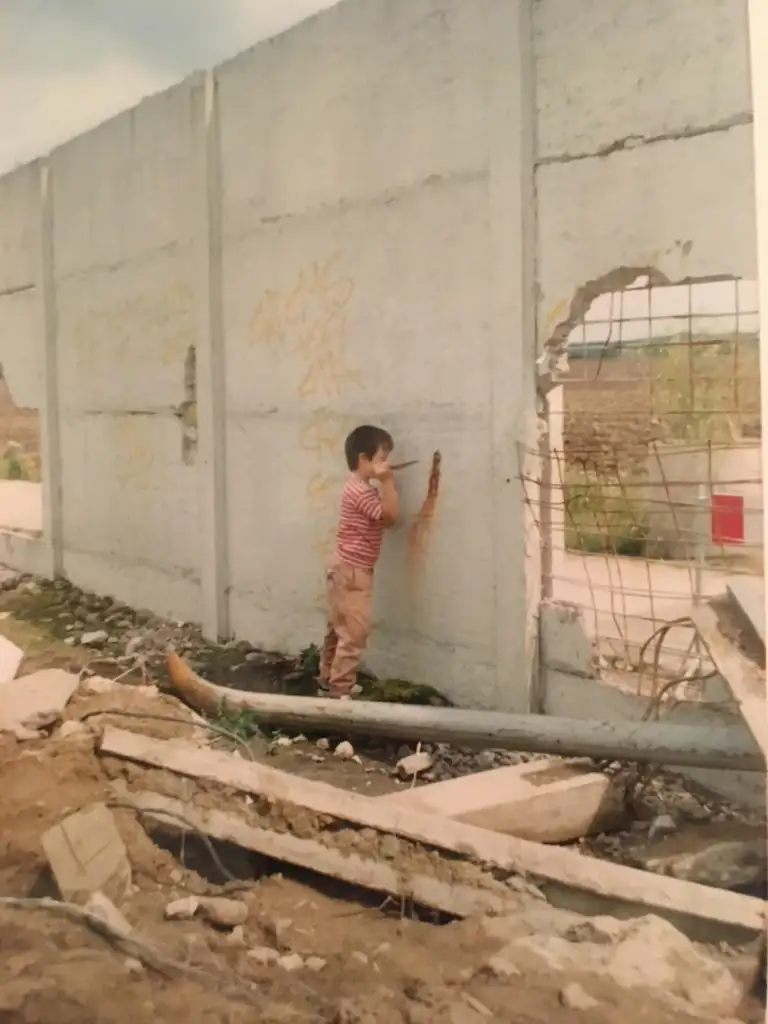 A child in a striped shirt and pants stands facing a damaged concrete wall with exposed rebar. The ground is littered with debris. The wall has graffiti, and there is an open area visible through the broken section. The sky is overcast.