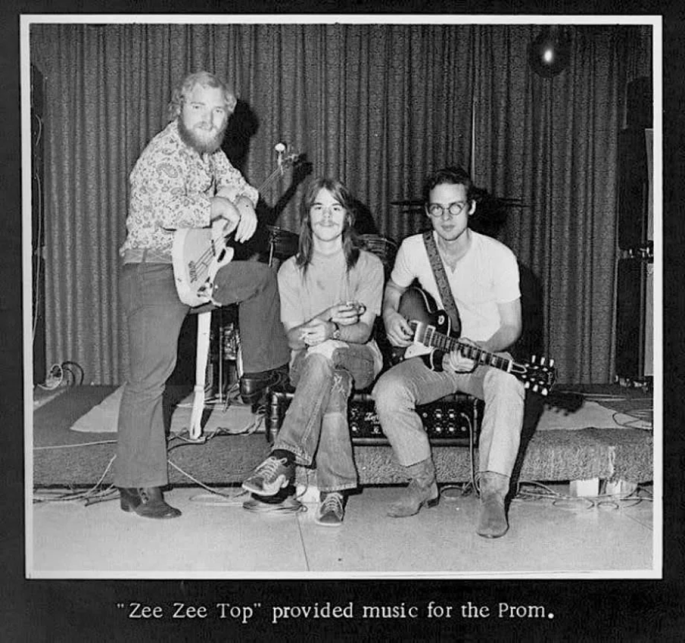 Three musicians pose with instruments on stage, with two seated and one standing. The standing member holds a bass guitar, while the others hold electric guitars. A drum set is visible behind them. Caption reads: "Zee Zee Top" provided music for the Prom.