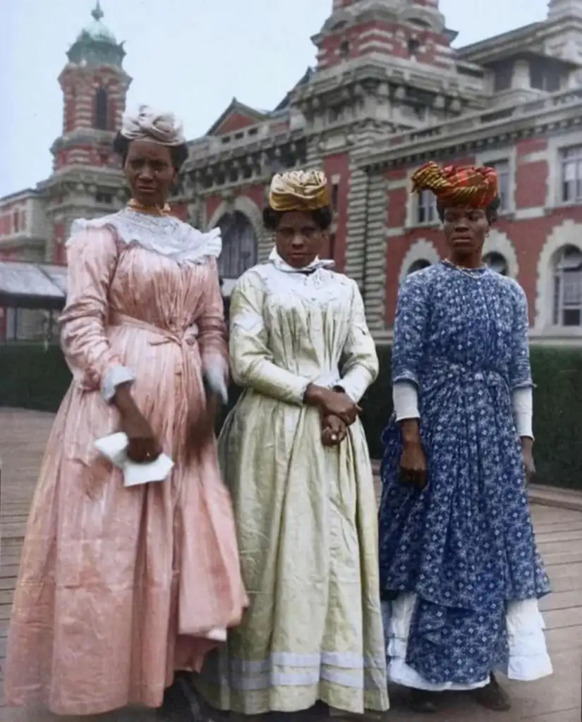 Three women in colorful, long dresses and headwraps stand in front of a historic brick building with intricate architecture.