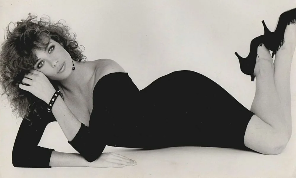 Woman with curly hair in a form-fitting black dress and high heels, lying on her side with legs bent and head propped up by her hand. The image is in black and white.