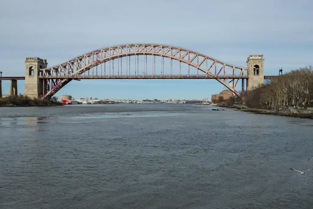 Arched steel bridge with two stone towers spanning a wide river, surrounded by bare trees on the right and an expanse of water beneath. A few ships are visible in the distance under a cloudy sky.