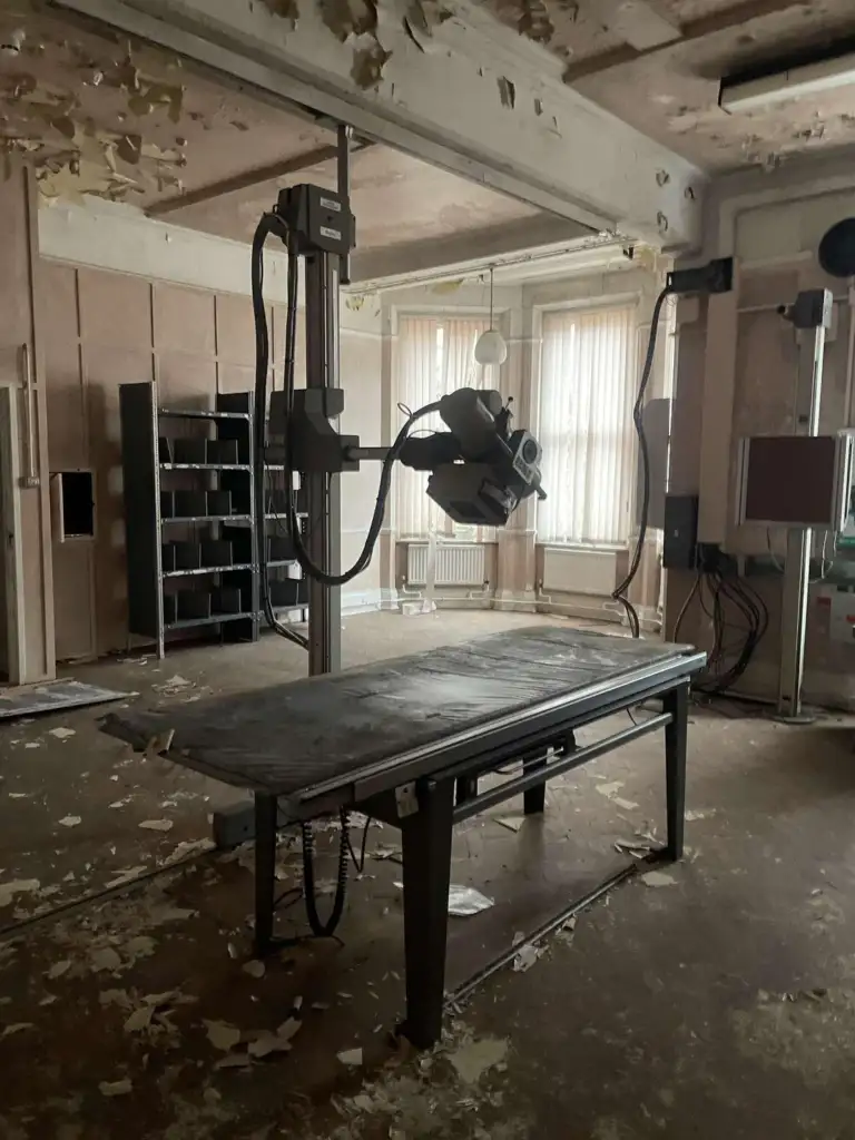 A dilapidated room with peeling paint and debris on the floor, featuring an old X-ray machine and table. Sunlight filters through dusty windows, highlighting the neglected state of the space.