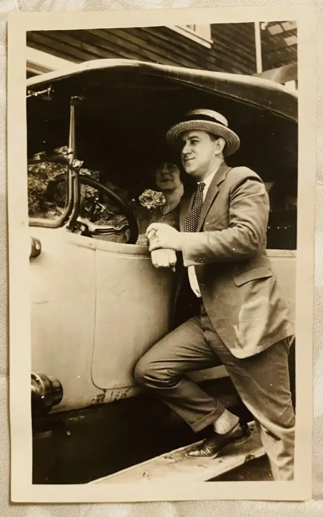 A vintage black-and-white photo shows a smiling man in a suit and straw hat standing next to an old car with his foot on the running board. A woman wearing a hat sits inside the car, partially visible through the open window.