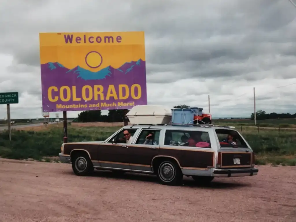 A vintage station wagon with a roof cargo box and visible luggage inside is parked next to a "Welcome to Colorado" sign. The sky is overcast, and the surroundings are grassy with a small sign for Sedgwick County.