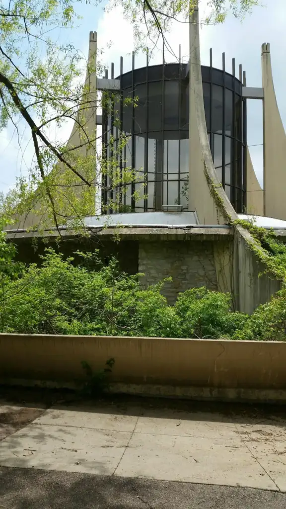 A modern building with a circular glass tower and tall, narrow concrete supports surrounded by lush green foliage. A low concrete wall runs in front, with a tree's branches overhanging from above.