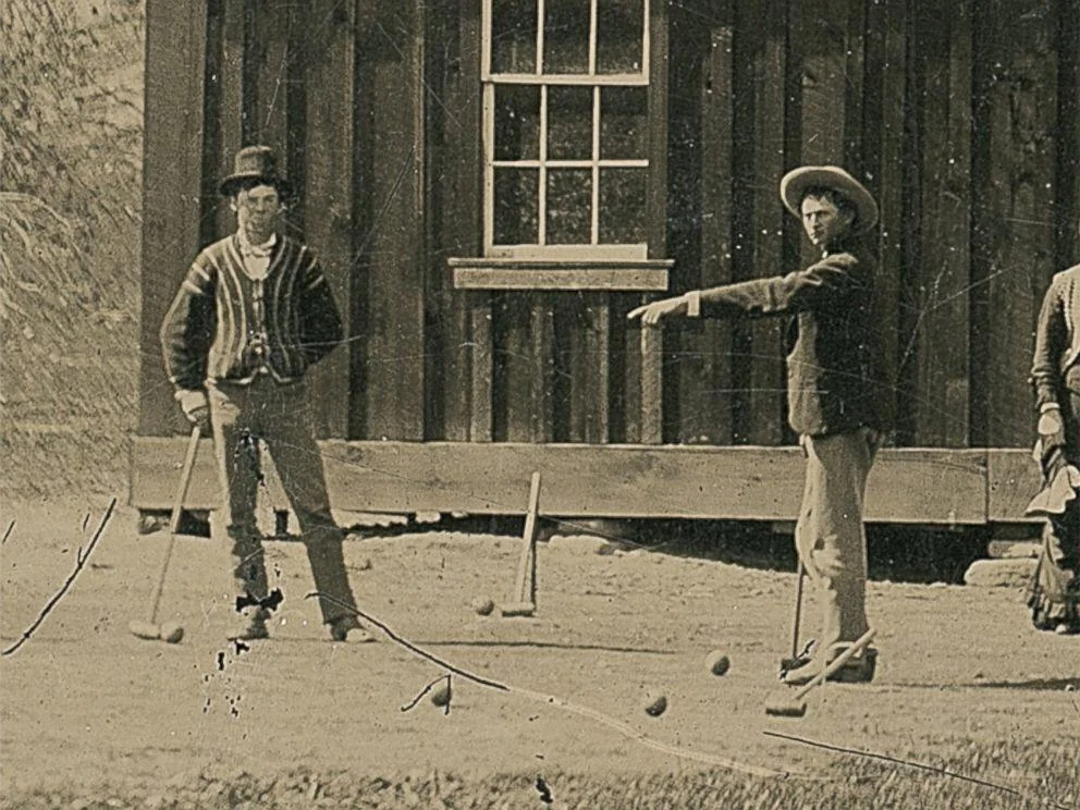 Two men playing croquet stand on grass in front of a wooden building. One man points while the other holds a croquet mallet. Both wear hats and period clothing, likely from the late 19th or early 20th century. A third person is partially visible on the right.