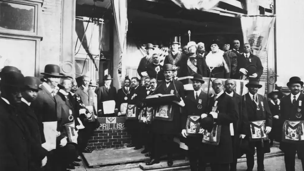 A historical black and white photo of a large group of men in suits and hats, standing on steps outside a building. They hold various objects, and a brick shows the inscription "April 1912." Several banners hang in the background.