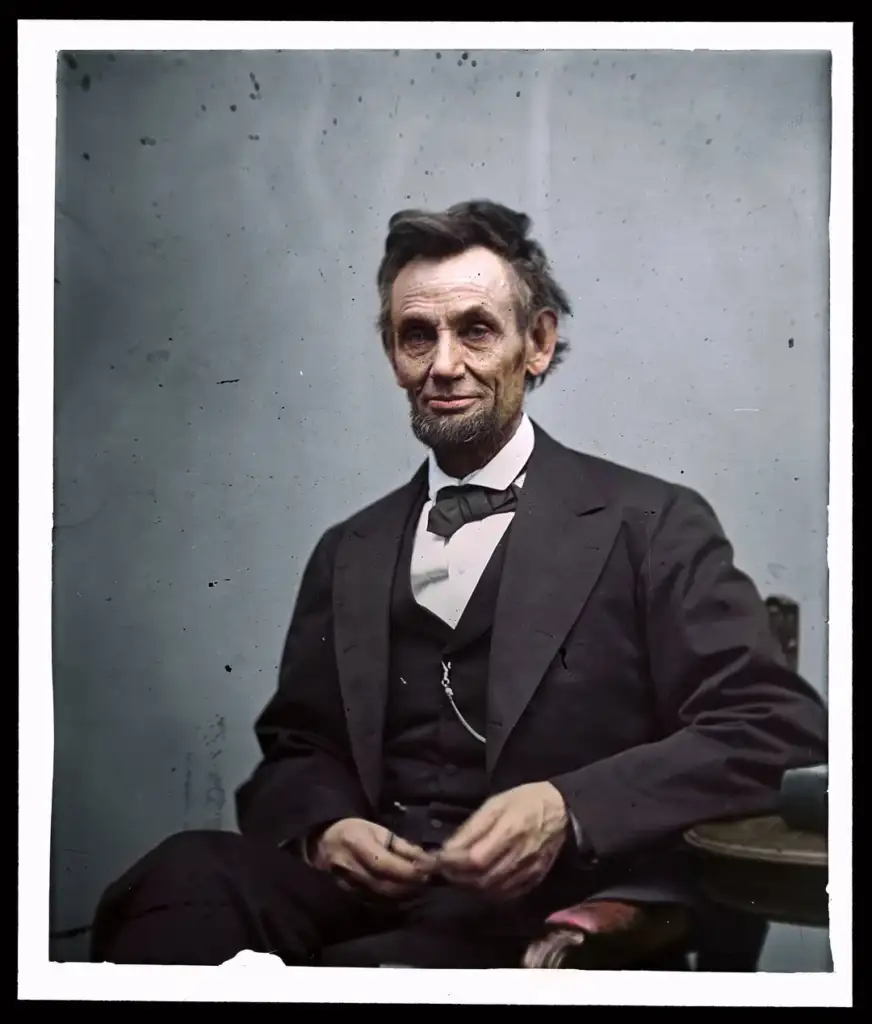 A historical figure with a beard and unkempt hair sits in a chair wearing a dark suit, waistcoat, and bowtie. He has a serious expression and holds his hands together in his lap, against a plain backdrop.