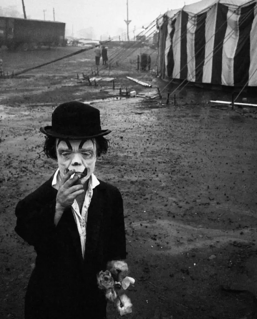 A clown smoking a cigarette stands in a muddy area near a striped circus tent. The clown wears a black bowler hat and holds flowers, with face paint giving a somber expression. The background is misty with faint figures and a gloomy ambiance.