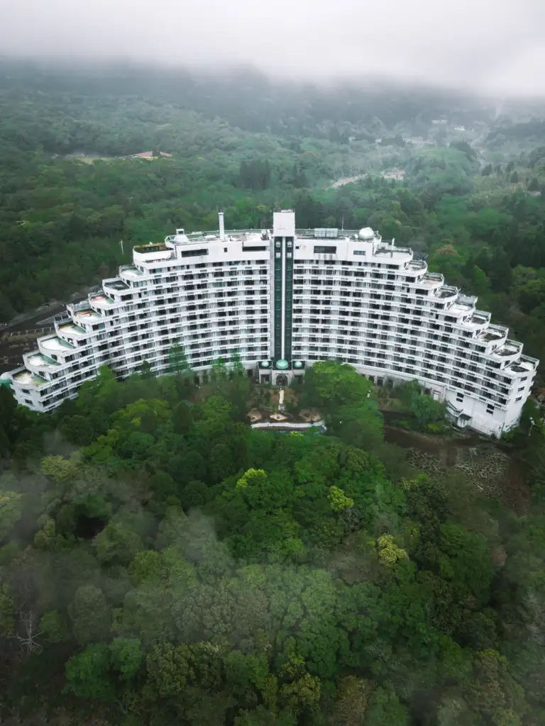 Aerial view of a large, curved white hotel surrounded by lush green forest. The building has many balconies and is nestled in a foggy, mountainous landscape, enhancing its secluded appearance.