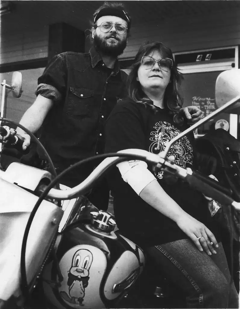 A man and woman pose confidently next to a motorcycle outside a building. The motorcycle features a cartoon duck on the fuel tank. Both are wearing glasses and casual clothing. The man has a beard and cap, while the woman wears a patterned shirt.