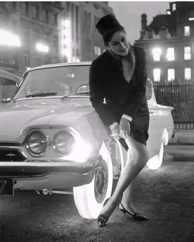 Black and white photo of a woman in a dark coat and heels, leaning against a classic car with glowing white tires. The background features a dimly lit street scene with city buildings.